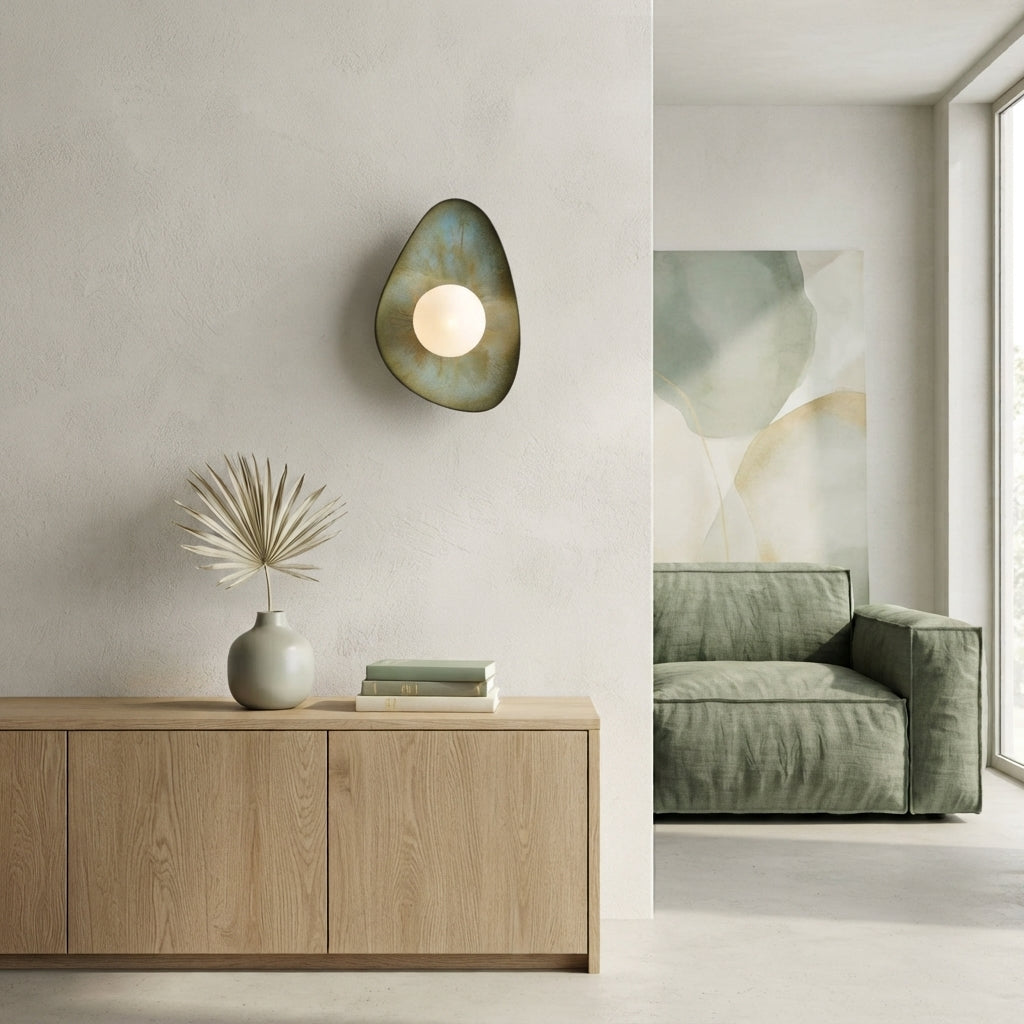 Handmade ceramic wall sconce in moss green, soft white, earthy brown, and a subtle hint of sky blue, featuring an organic sculptural shape inspired by smooth seashore pebbles. The ceramic surface has natural variations in colour and texture, with a round frosted glass globe that gently diffuses warm light. Handmade in Lithuania and designed for hardwired installation.