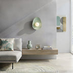 Handmade ceramic wall sconce in soft jade pastel green, featuring an organic sculptural shape inspired by smooth seashore pebbles. The stoneware has a smooth matte surface with subtle variations in colour and texture. A round frosted glass globe sits in the center, gently diffusing warm light. Handmade in Lithuania and designed for hardwired installation.